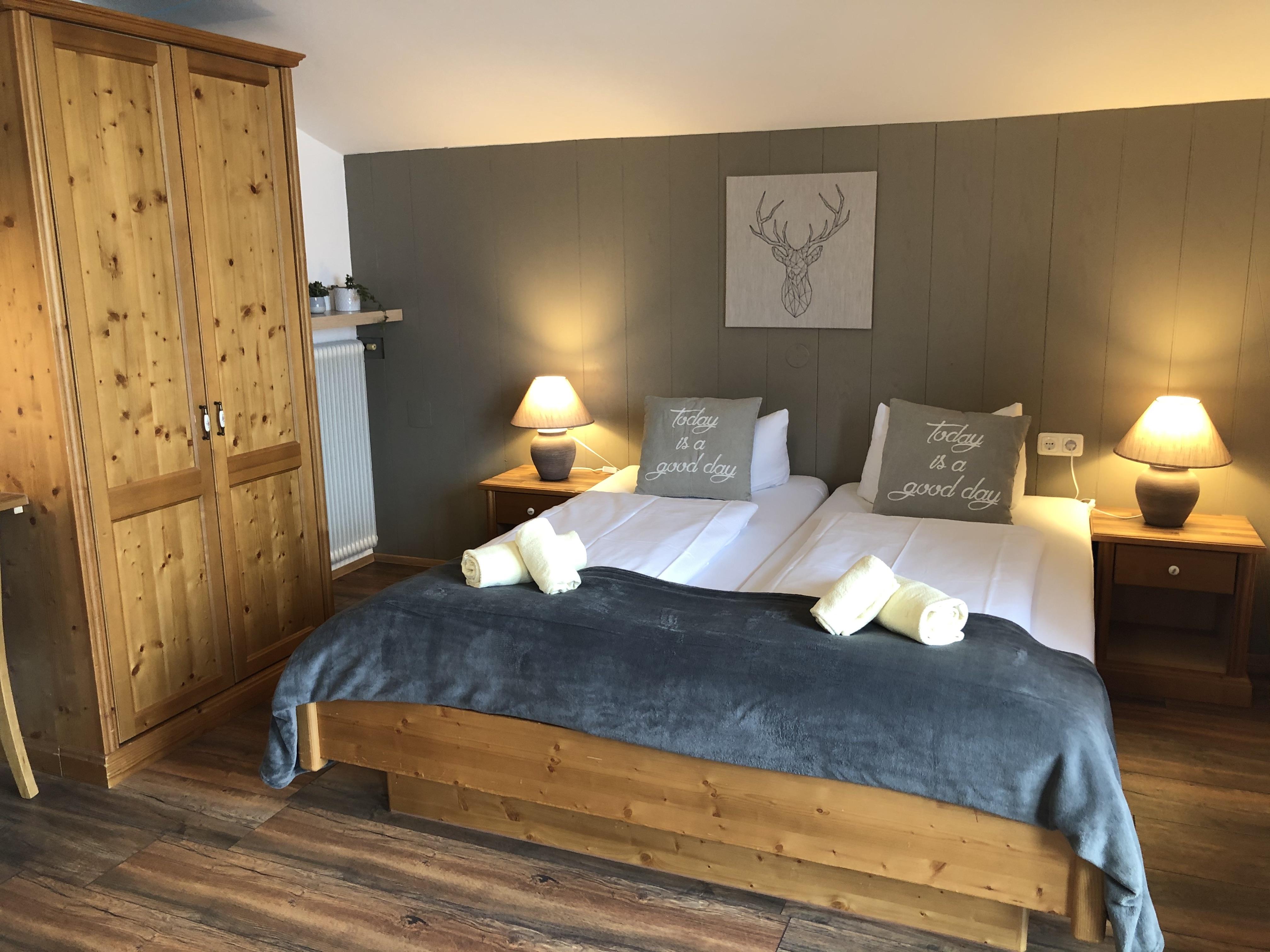A cozy bedroom with two beds, nightstands, and warm light. The wooden furniture and decoration create an inviting atmosphere.