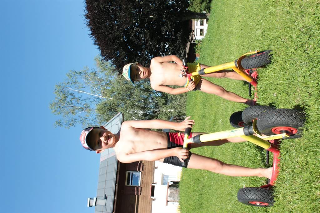Two boys are standing outside on large scooters. They are wearing swimwear and caps and enjoying the sunny day.