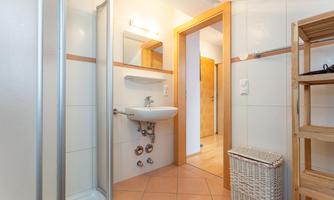 A modern bathroom with a shower, a sink, and bright tiles. There is also a laundry basket and a wooden shelf.