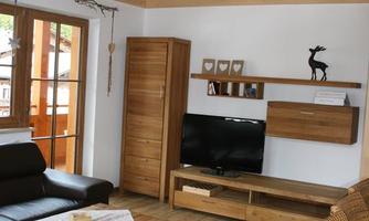 A modern living room with wooden flooring and large windows. There is a comfortable seating set, a TV, and an open roof area above.