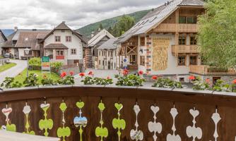 A balcony with rustic wood design offers a view of a picturesque village. The surroundings are characterized by green forests and traditional houses.