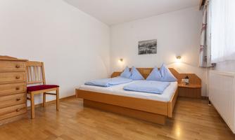A simple bedroom with a double bed and two pillows. There is a wooden desk and a chair as well as a bright, friendly room with wooden flooring.