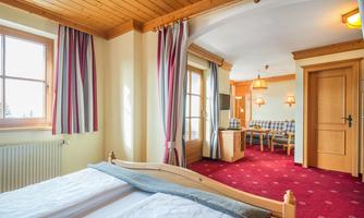 A cozy hotel room with wooden walls and red carpets. There is a double bed, windows with curtains, and a seating area.