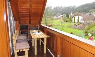 A cozy balcony with a wood-paneled wall and a small table. In the background, there are green meadows and a picturesque village.