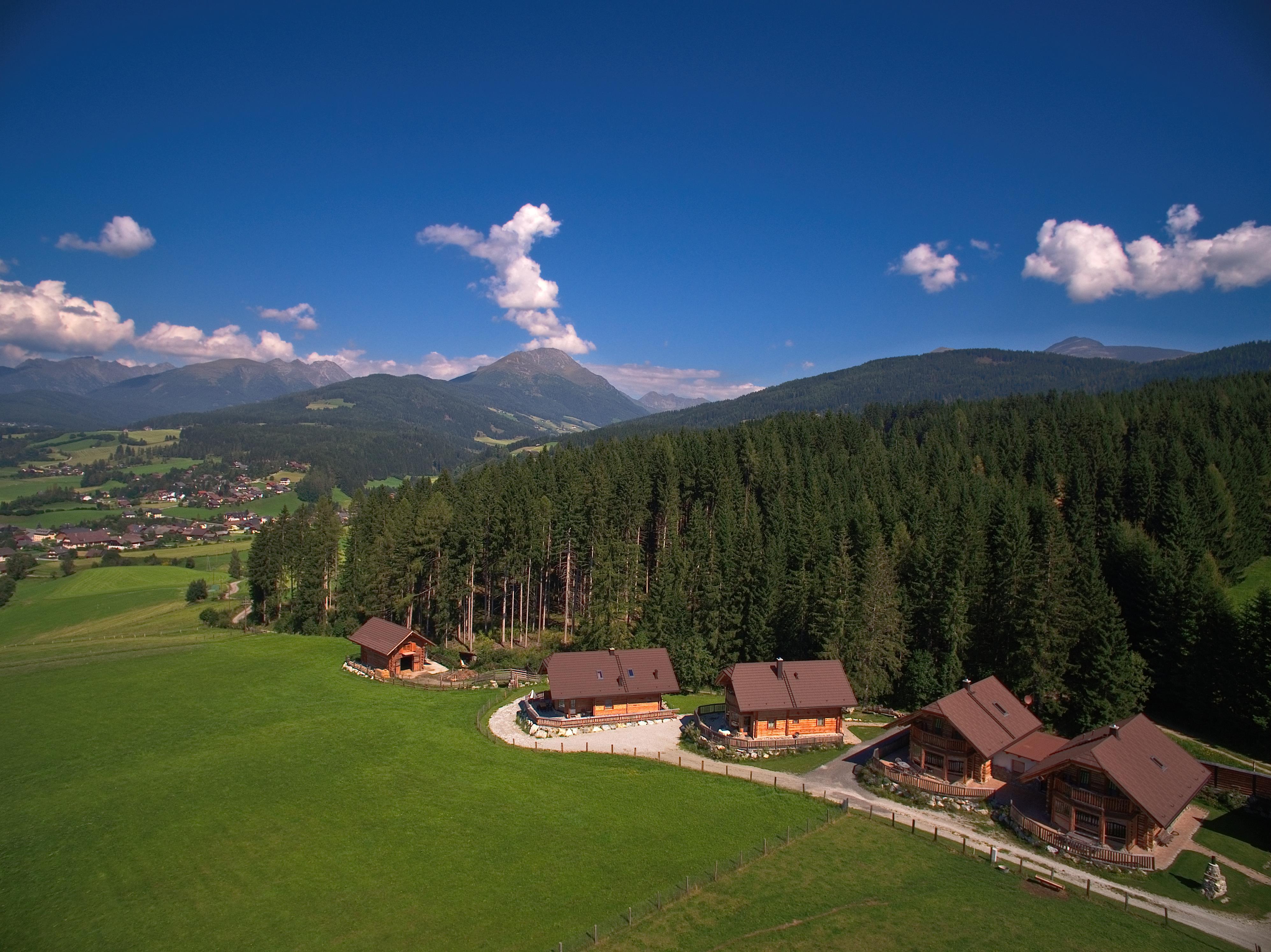 A picturesque landscape with green meadows and dense forests. In the foreground, several cozy cabins stand in front of an impressive mountain backdrop.