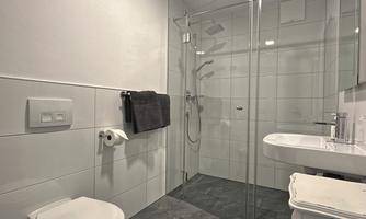 A modern bathroom with a glass shower and bright, minimalist design. The walls are tiled in white and the floor is in dark stone.