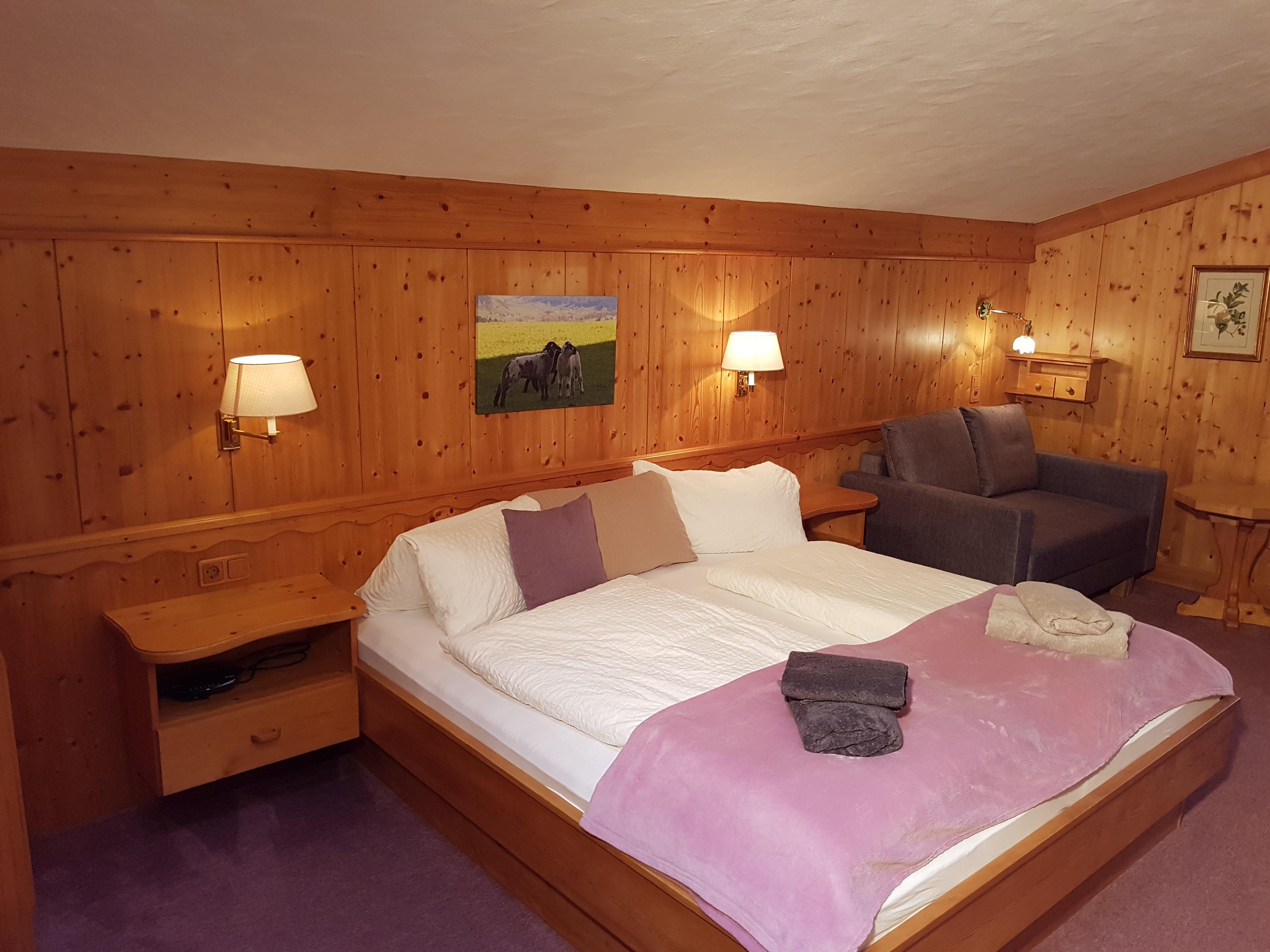 A cozy bedroom with wood paneling, a large bed, and a sitting area. The decoration is simple, with soft colors and artificial light.