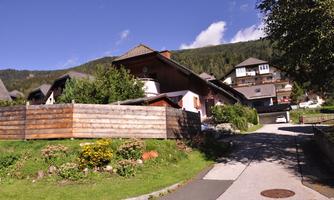 A picturesque street with traditional houses and green gardens. In the background, gentle hills can be seen.