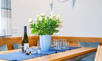 A stylishly set table with a blue tablecloth, a bottle of wine, and a vase with white roses. In the background, cozy seat cushions can be seen.