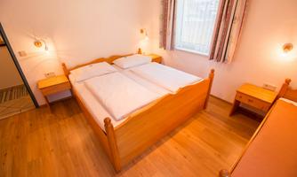 A simple bedroom with two single beds and wooden clocks. The room has a bright floor and large windows.