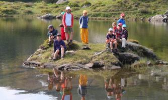 A group of children is standing on a small rock in a calm lake. In the background, green hills and trees can be seen.