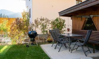 A cozy terrace with a table, chairs, and a grill. In the background, colorful plants and a calm environment can be seen.
