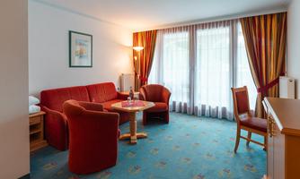 A cozy living room with a red sofa and a round table. Large windows with curtains let in a lot of light.