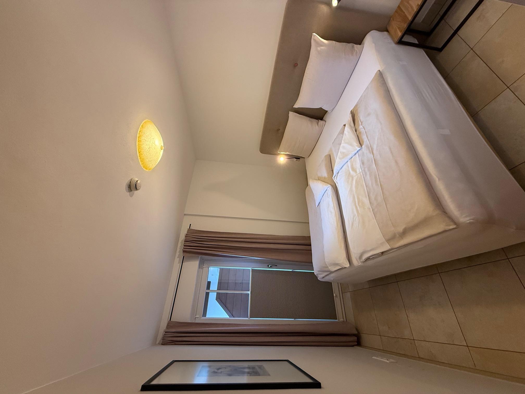 A cozy bedroom with two beds and a window that lets in daylight. The walls are bright, and the room feels modern and inviting.