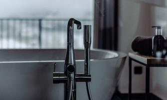 An elegant bathtub with a modern black faucet and a hand shower. In the background, there is a stylish table with care products.