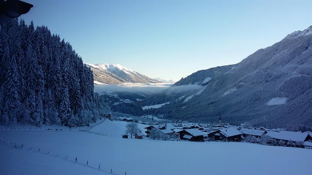 A snowy landscape with tall trees and mountain peaks in the background. In the valley, small huts can be seen, surrounded by snow-covered ground.