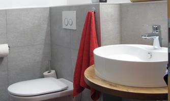 A modern bathroom with gray tiles and a round sink. A red towel rack and a red bath mat add color to the space.