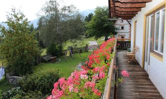 A balcony with colorful flowers and a view of a green landscape. In the background, there are trees and some buildings visible.