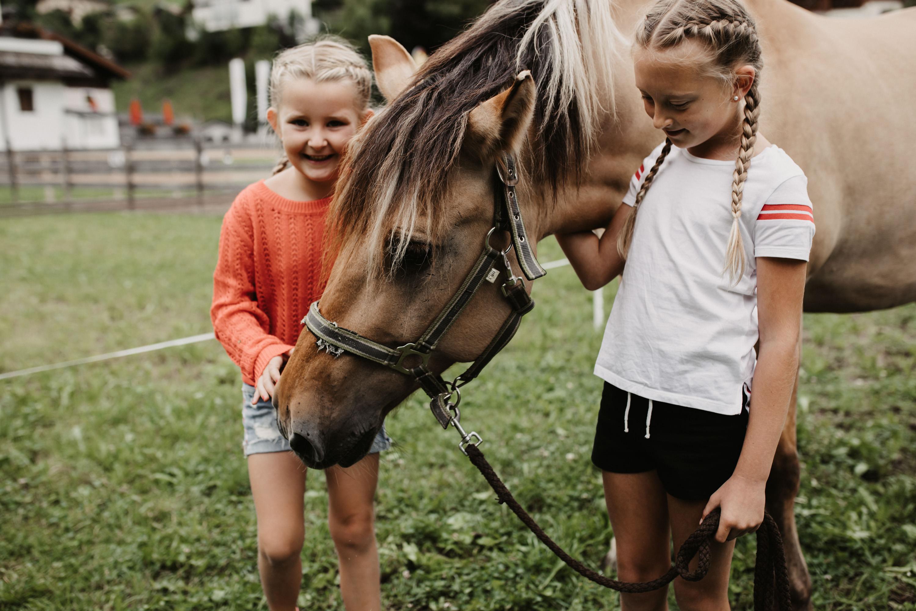 Two girls are standing in a meadow and are engaging with a horse. Both are smiling and seem to be having fun.