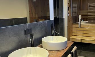 A modern bathroom with two round sinks on a wooden countertop. In the background, wooden paneling and a sauna are visible.