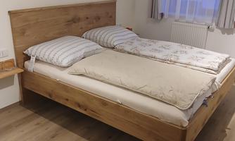 A cozy bedroom with a wooden double bed. The walls are light, and the windows let in a lot of light.