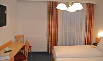 A simple hotel room with two beds, a table and chairs. Curtains in warm colors and a large window front create an inviting atmosphere.