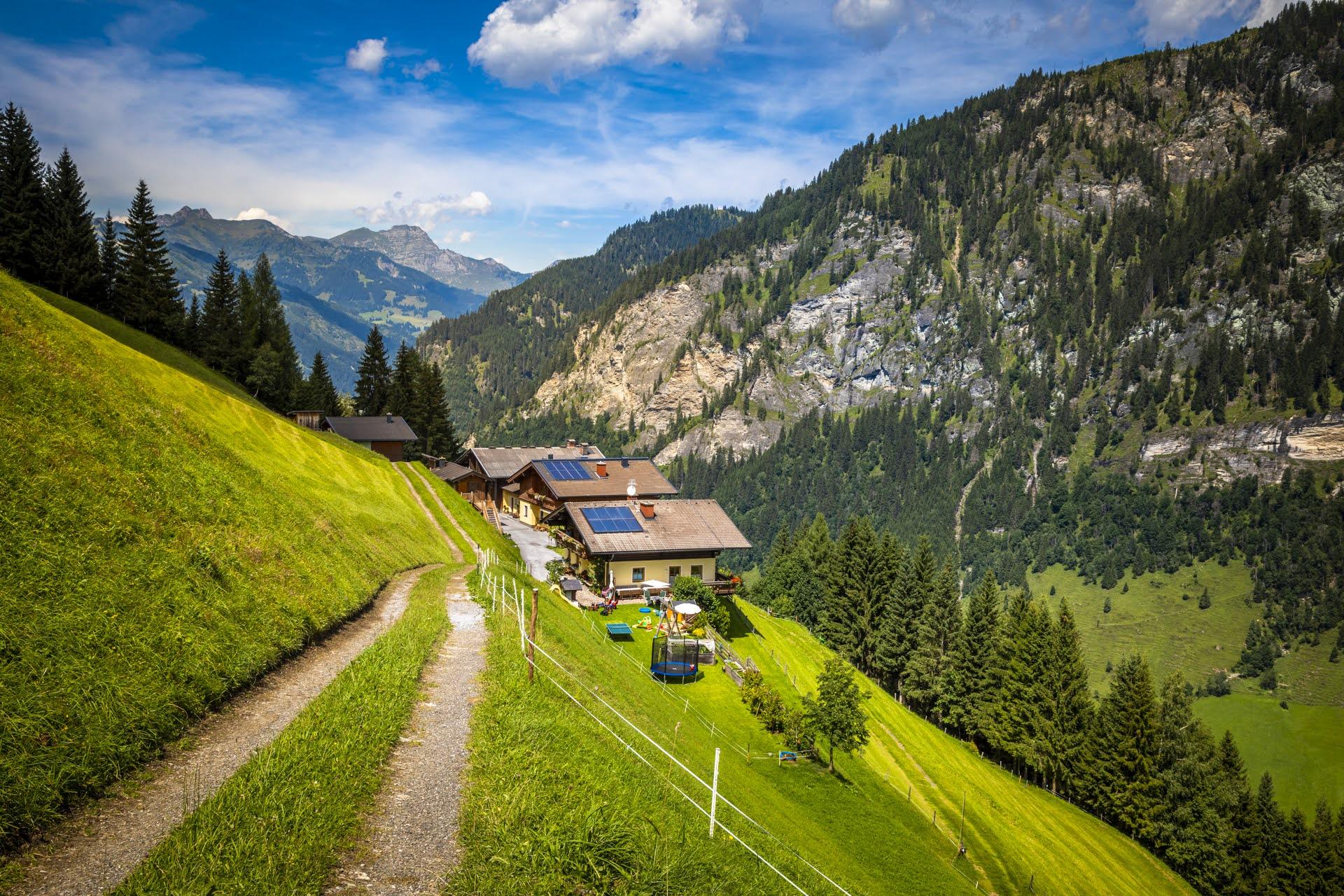 A picturesque mountain landscape with a traditional house and green meadows. In the background, majestic mountains rise beneath a clear sky.