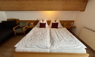 A cozy bedroom with a large double bed and pillows in white and burgundy. The room has a warm ambiance with wooden details and a comfortable armchair.