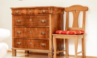 A classic wooden cabinet with three drawers stands next to a simple wooden chair. The chair has a red cushion and enhances the rustic charm of the room.