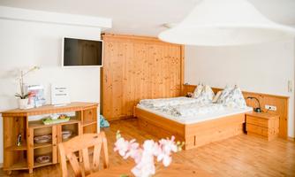 A cozy room with a double bed, wooden furniture, and a television. Fresh flowers are on the table, and the room is bright and welcoming.