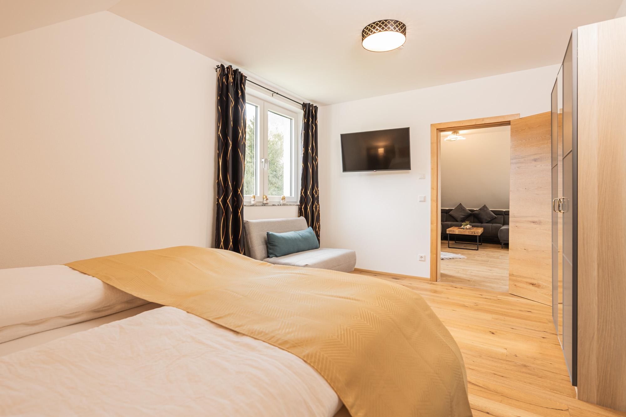 A modern bedroom with a double bed and a television on the wall. The room has large windows and a cozy seating area.