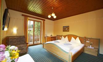 A cozy room with a double bed and a bright wooden ceiling. There is an armchair and a balcony with a view of nature.