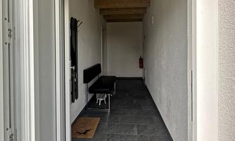 A bright corridor with a wooden ceiling and gray tiles. On the right side stands a bench and a small rug lies in front of the door.