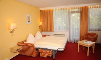 A cozy hotel room with orange curtains and a small table. The beds are neatly made and the room gets plenty of daylight.