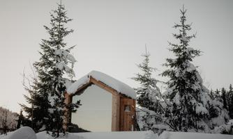 A wooden building with a large mirror is situated in a snowy landscape. Next to the building, tall, snow-covered fir trees grow.