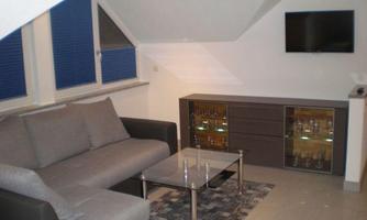 A modern living room with a gray couch, a glass table, and a television on the wall. Light is streaming in through the windows, creating a bright atmosphere.