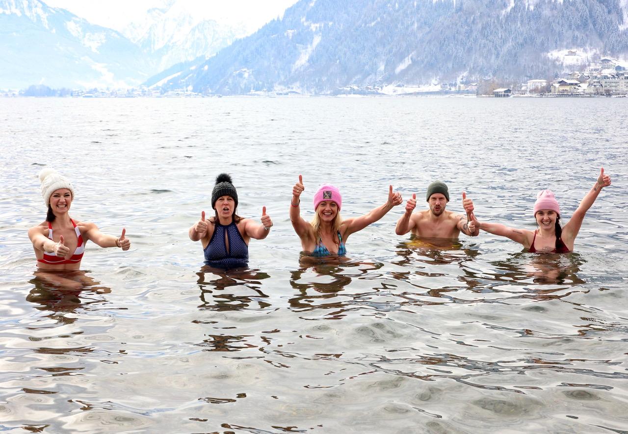 Five people are standing in the cold water and giving a thumbs up. They are wearing colorful hats and laughing happily.