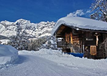 A snow-covered mountain hut surrounded by snow-covered trees and majestic mountains. The sky is clear and blue.