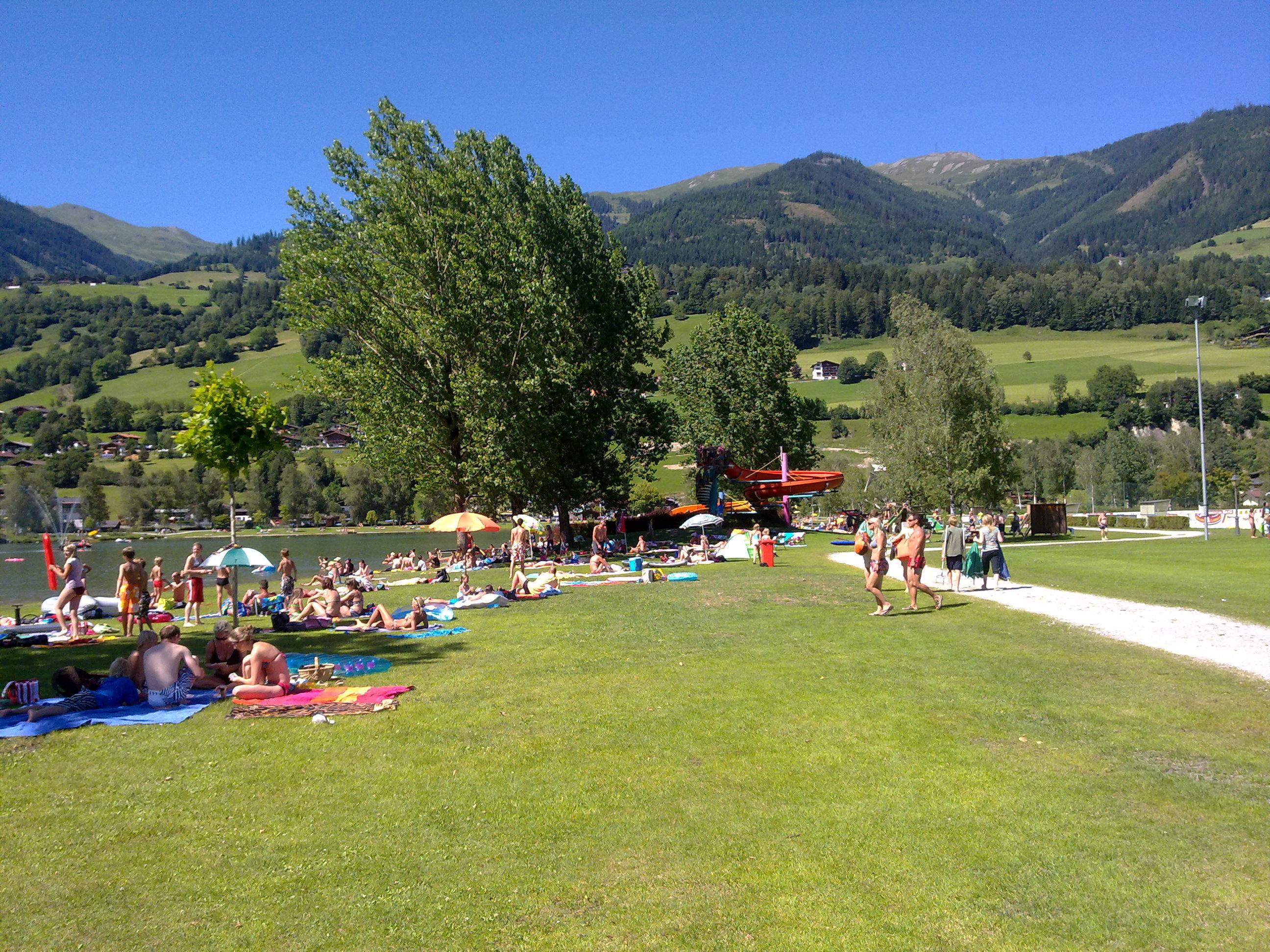 A lively park by the water with many people relaxing on blankets. In the background, green hills and trees can be seen under a blue sky.
