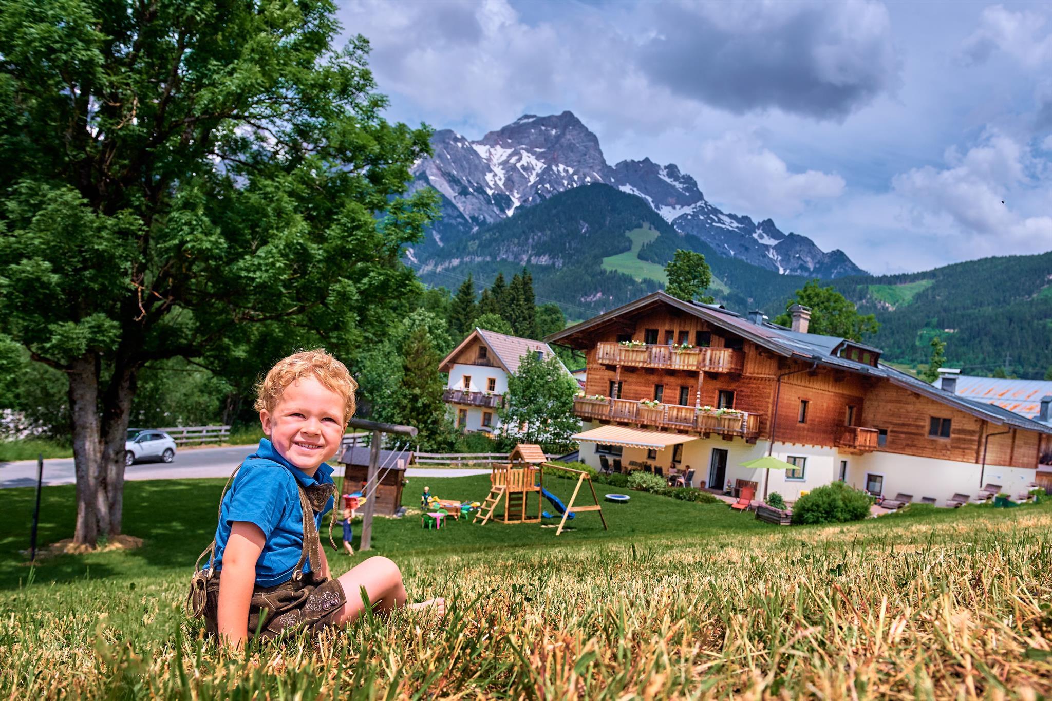 A happy child sits on a meadow in front of a beautiful wooden house. In the background, mountains and a playground can be seen.
