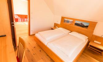 A bright bedroom with a double bed and a wooden frame. In the background, a living room with a red sofa can be seen.