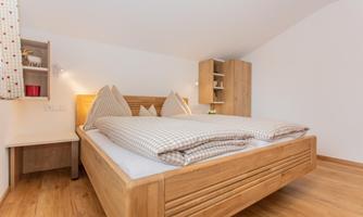 A modern bedroom with a large wooden bed and two cushions. The walls are bright and the floor is made of wood.