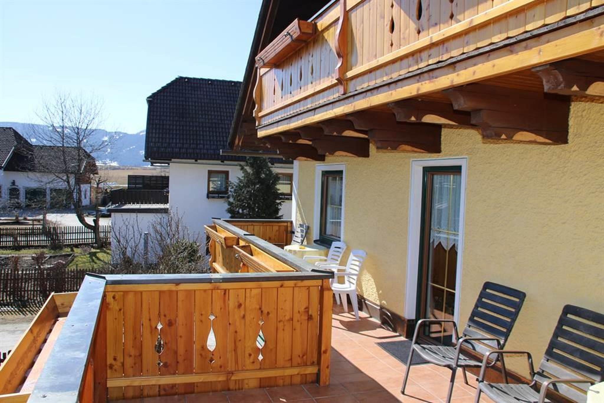 A cozy balcony with a wooden railing and deck chairs. In the background, houses and trees can be seen against a clear sky.