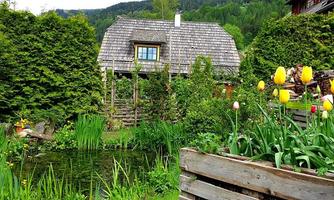 A picturesque garden with colorful tulips and a small pond. In the background stands a cozy wooden house, surrounded by green hedges and mountains.