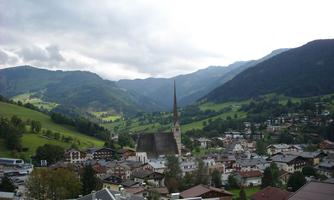 A picturesque landscape with a small village and a church in the foreground. In the background, green hills and mountains can be seen under a cloudy sky.