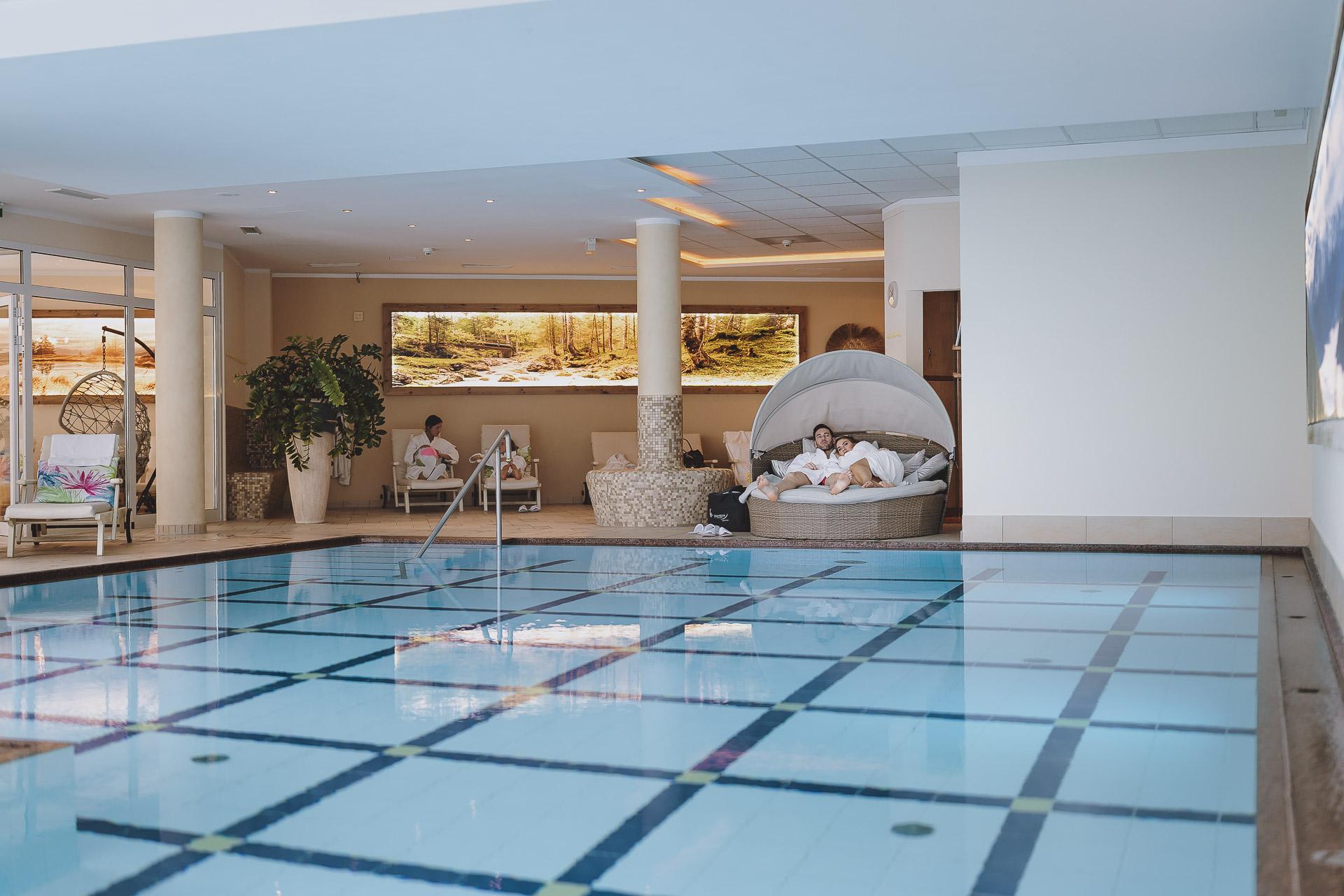 An indoor pool with clear water and a modern lounge area. The room is bright and inviting, with plants and comfortable furniture.