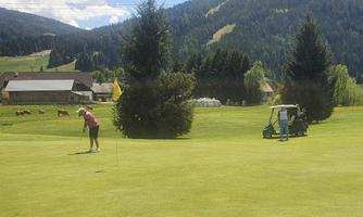 A golfer is putting on a green course with a view of the mountains. In the background, there are trees and a golf cart visible.