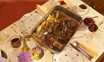 A beautifully set table with a tray full of meat and potatoes. Served alongside are red wine and a dessert.