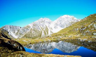 An impressive mountain landscape with snow-capped peaks and clear blue skies. In the foreground, the landscape is reflected in a calm lake.