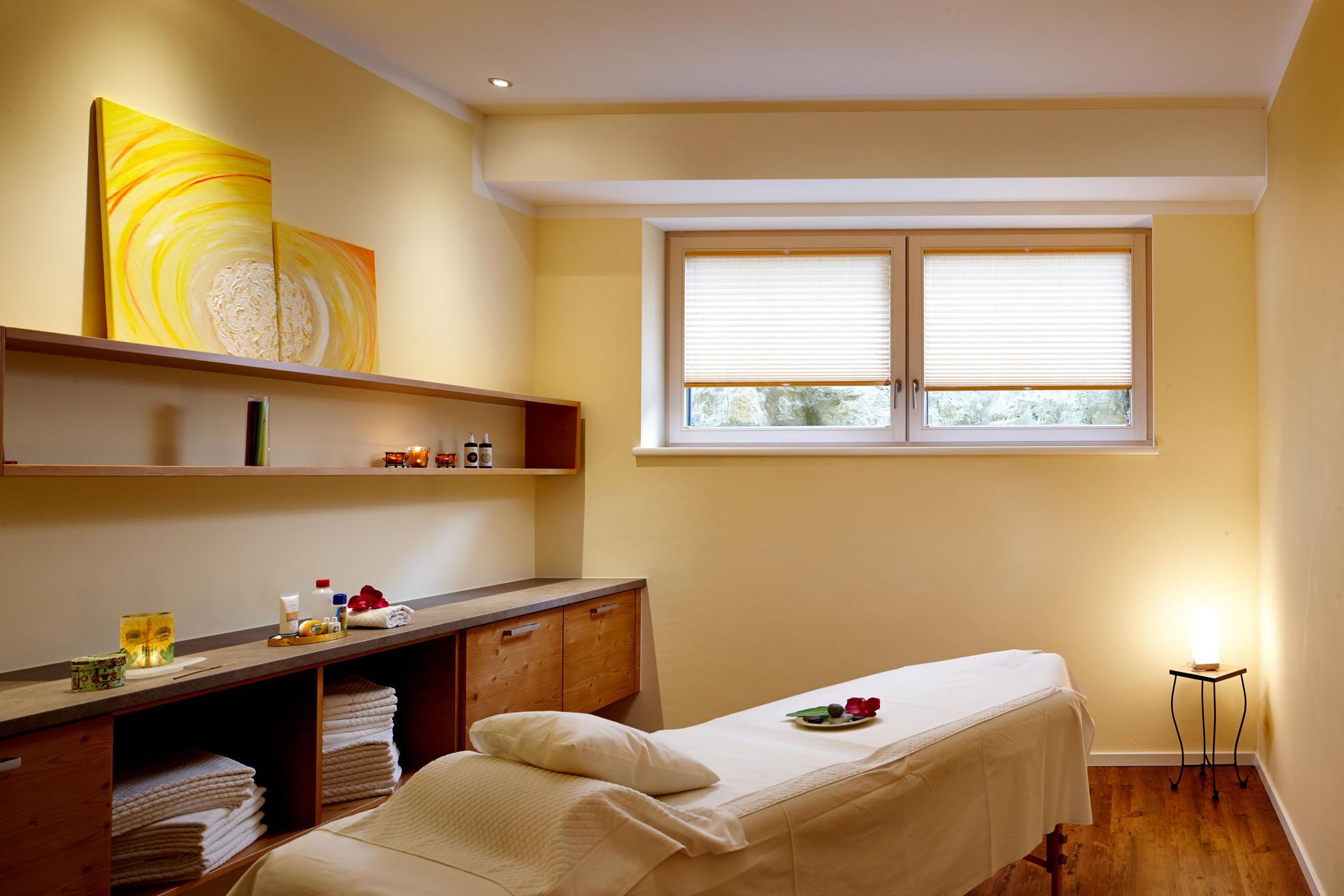 A relaxing wellness room with a massage table and soft colors. The walls are painted yellow, and there are windows overlooking the outdoors.